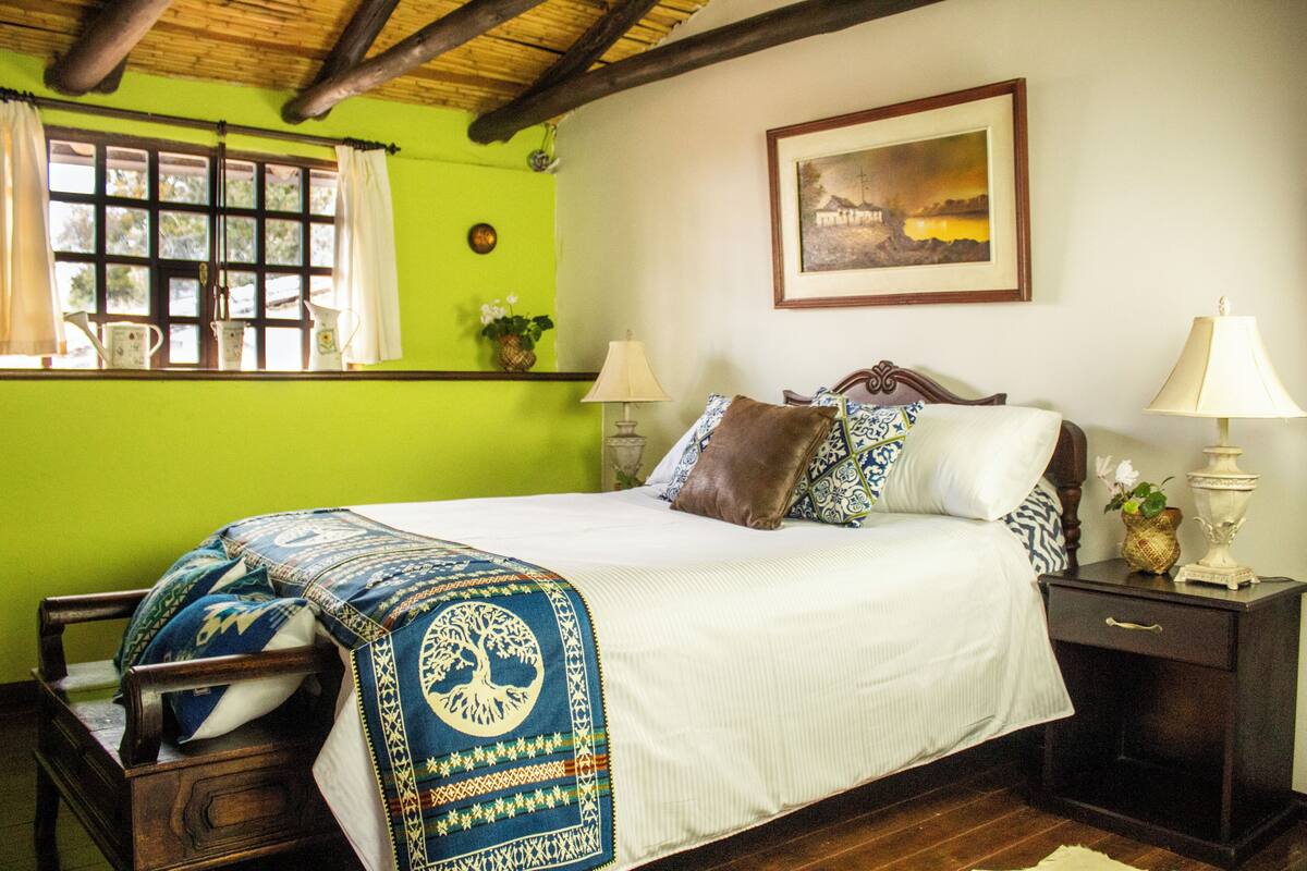 Un lit bien fait, recouvert d’une couverture à motifs bleus et blancs, une table de chevet en bois avec une lampe, et un tableau encadré accroché au mur.