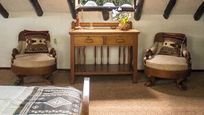 Una habitación con techo inclinado, vigas de madera, un ventanal grande, dos sillones, un escritorio de madera y una cama con ropa de cama estampada.