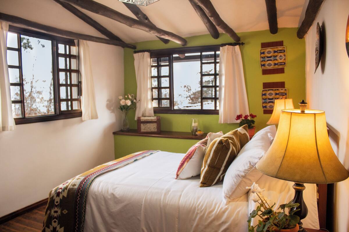 Une chambre à coucher comprenant un lit, une lampe de chevet, une fenêtre avec des rideaux, une petite table sur laquelle est posé un vase de fleurs, et un mur décoré d’assiettes.