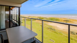 Un balcon avec une table et des chaises, donnant sur une plage bordée de dunes et l’océan.