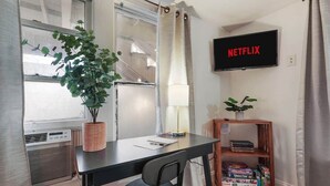 Ein kleines Zimmer mit Schreibtisch, Stuhl, Topfpflanze, Lampe, Fernseher und Bücherregal.