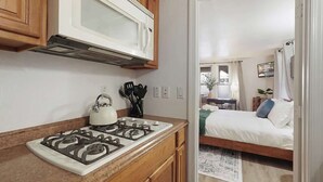 Eine kompakte Küche mit Gasherd, Holzschränken und Mikrowelle. Durch eine offene Tür ist ein Schlafzimmer mit Bett und Vorhängen zu sehen.