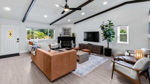 A modern living room with a brown sofa, a coffee table, and a TV. There is a fireplace, a large window, and a ceiling fan. - 3-bedroom Fireplace Stunning Spanish Home (Indio)