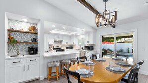 A modern kitchen with white cabinetry, a central island, and a dining area with a wooden table and black chairs. - 3-bedroom Fireplace Stunning Spanish Home (Indio)