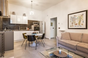 Uma cozinha moderna com armários brancos, uma sala de jantar com mesa redonda e cadeiras, e um sofá bege em uma sala de estar.