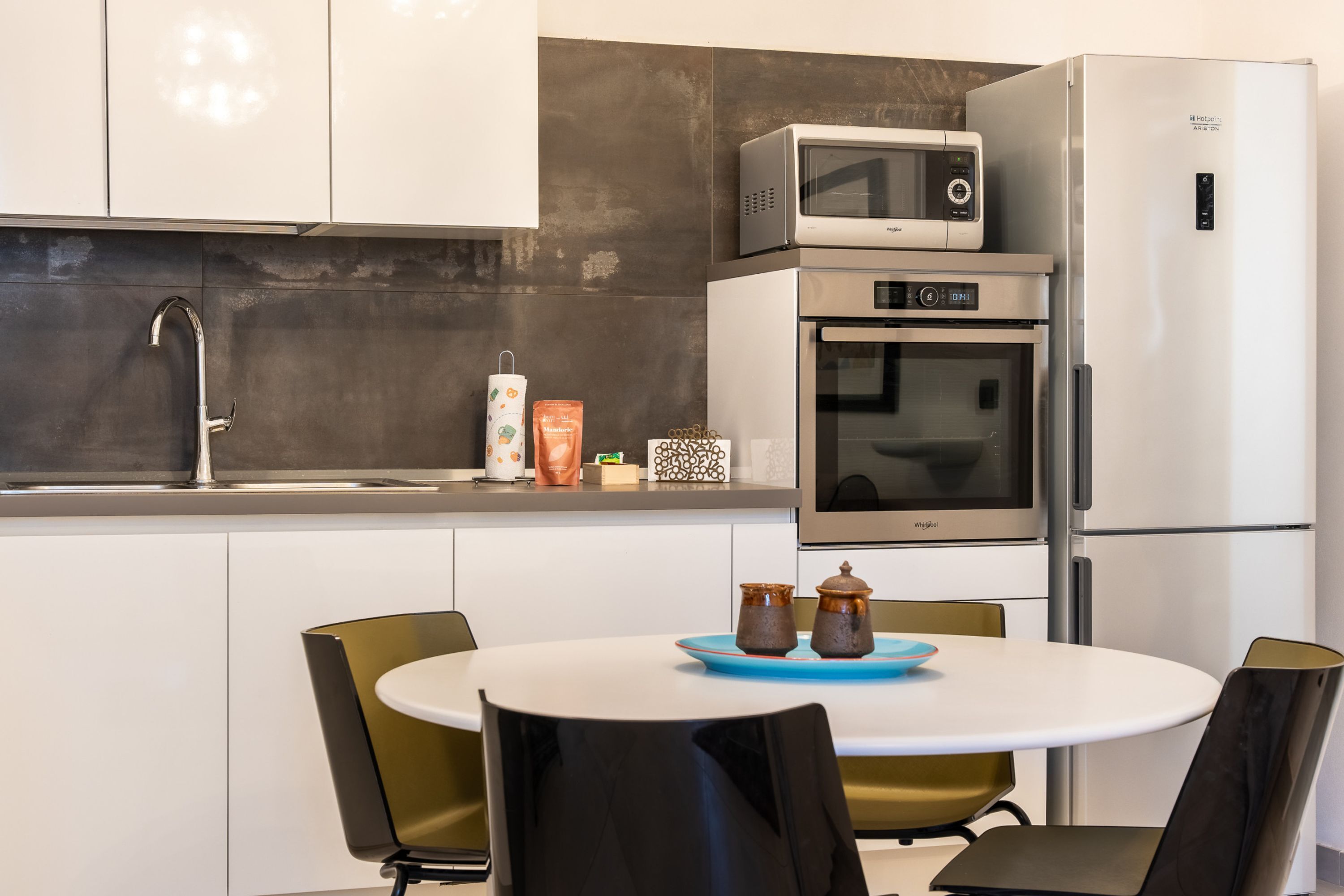 Una cocina moderna con microondas, horno y refrigerador. Hay un lavamanos con grifo, una mesa con sillas, y una bandeja azul con dos frascos.