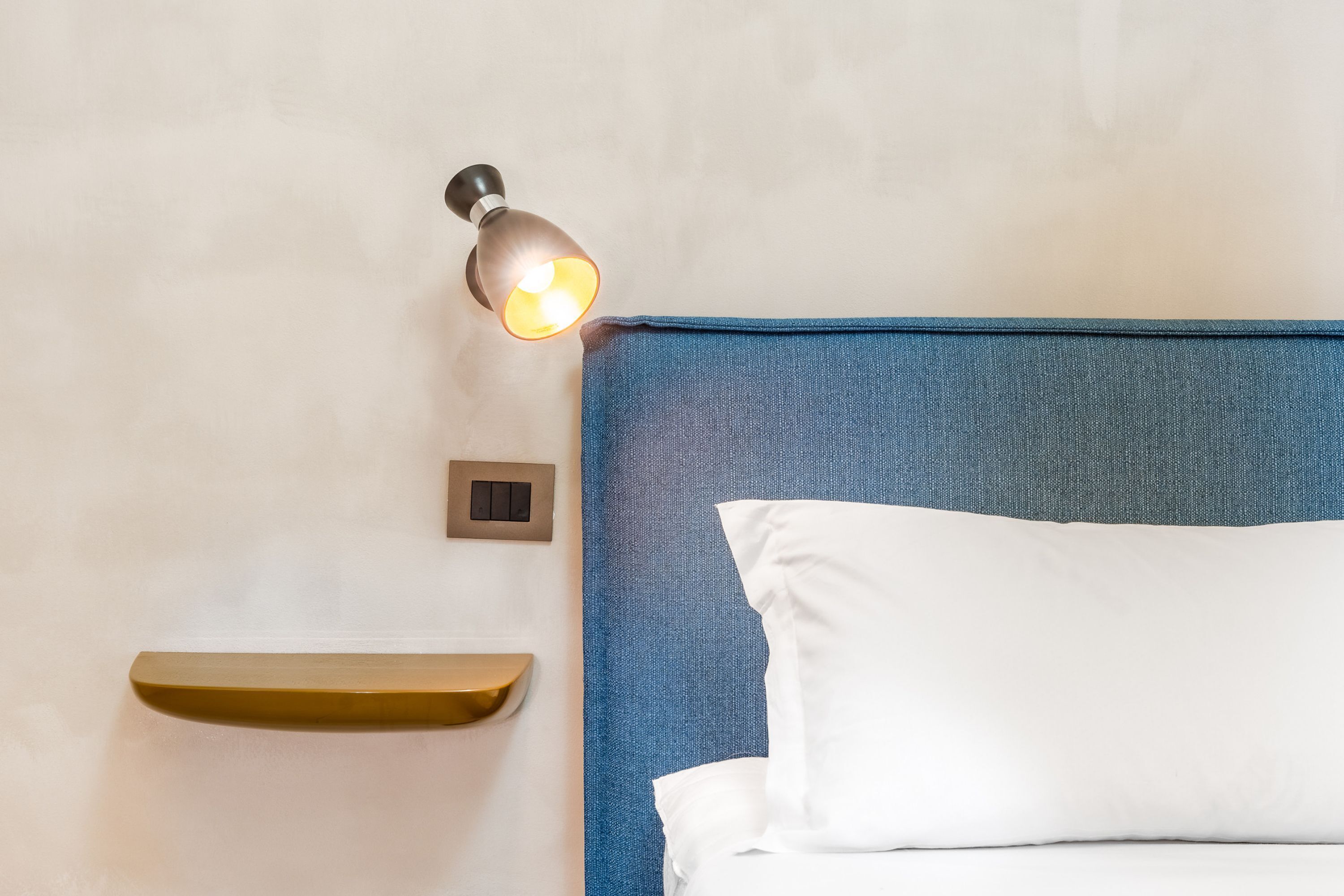 Una cama bien hecha con cabecera azul, una lámpara de noche y una pequeña estantería.