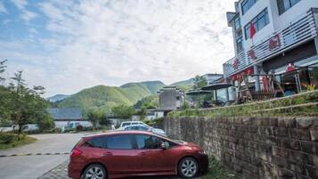 A red car parked on a paved area next to a stone wall, with a building featuring balconies and outdoor seating in the background.