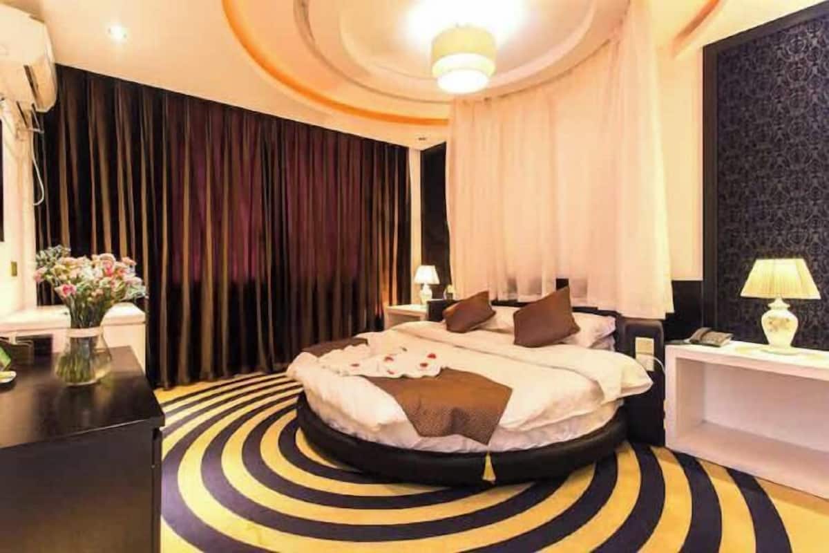A modern hotel room with a circular bed, dark curtains, a round rug, and a nightstand with a lamp.