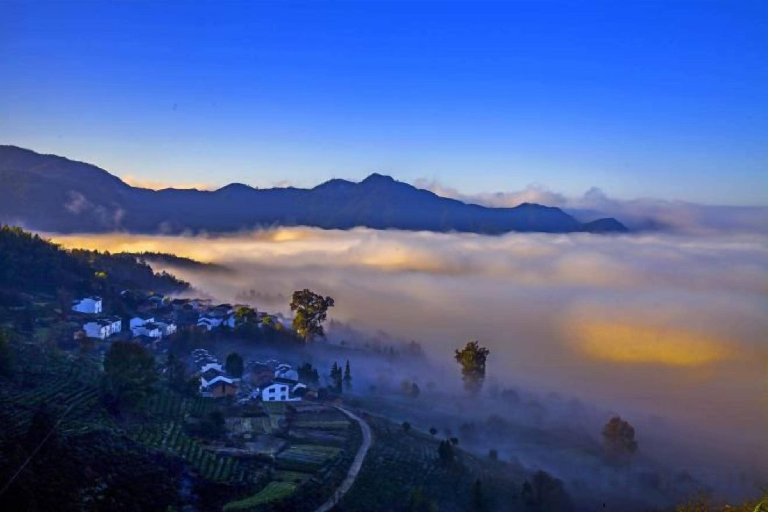 A village nestled in a valley with a clear sky above and a mountain range in the background.