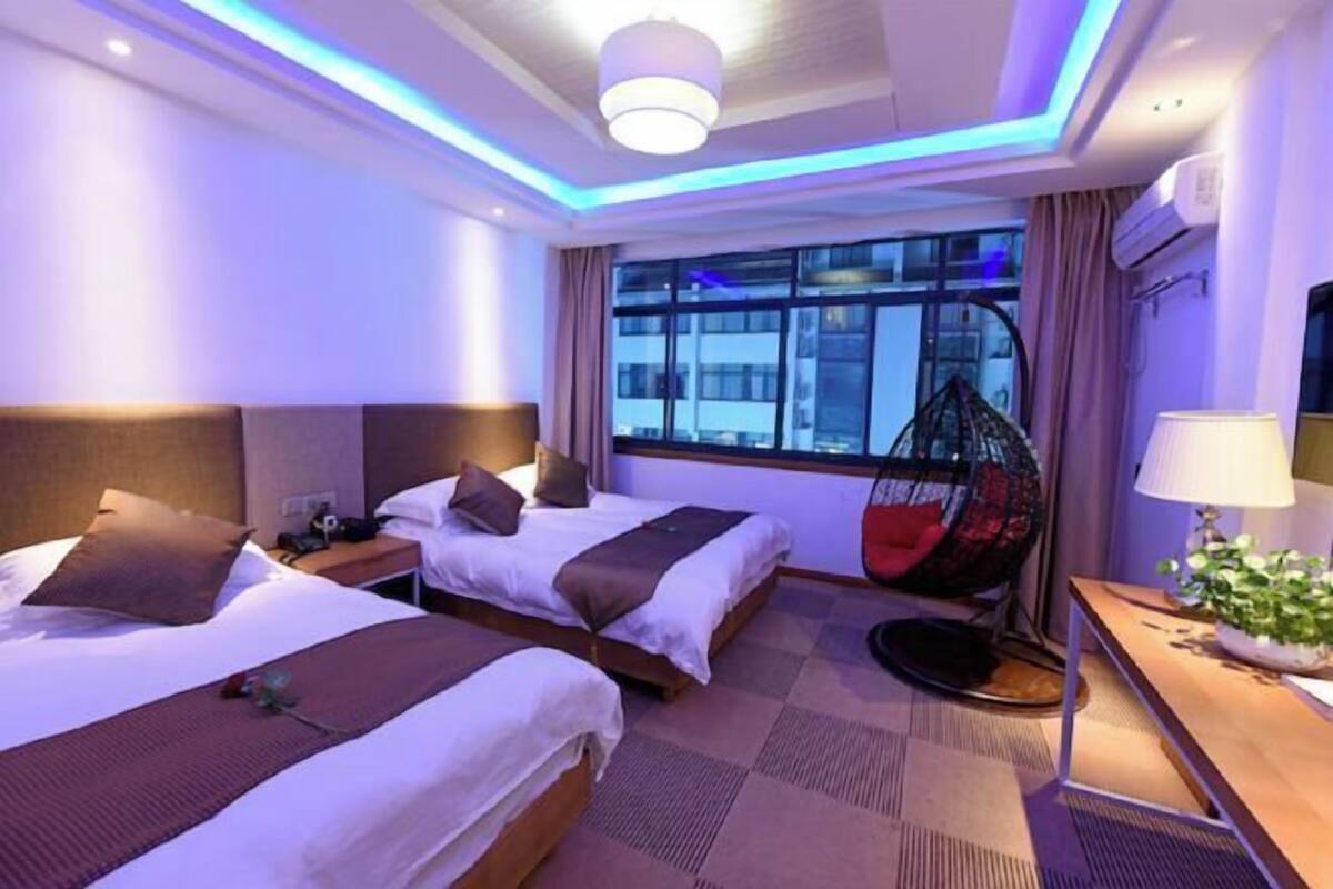 A hotel room with two beds, a hanging chair, a desk with a lamp, and a large window with a view of the city.
