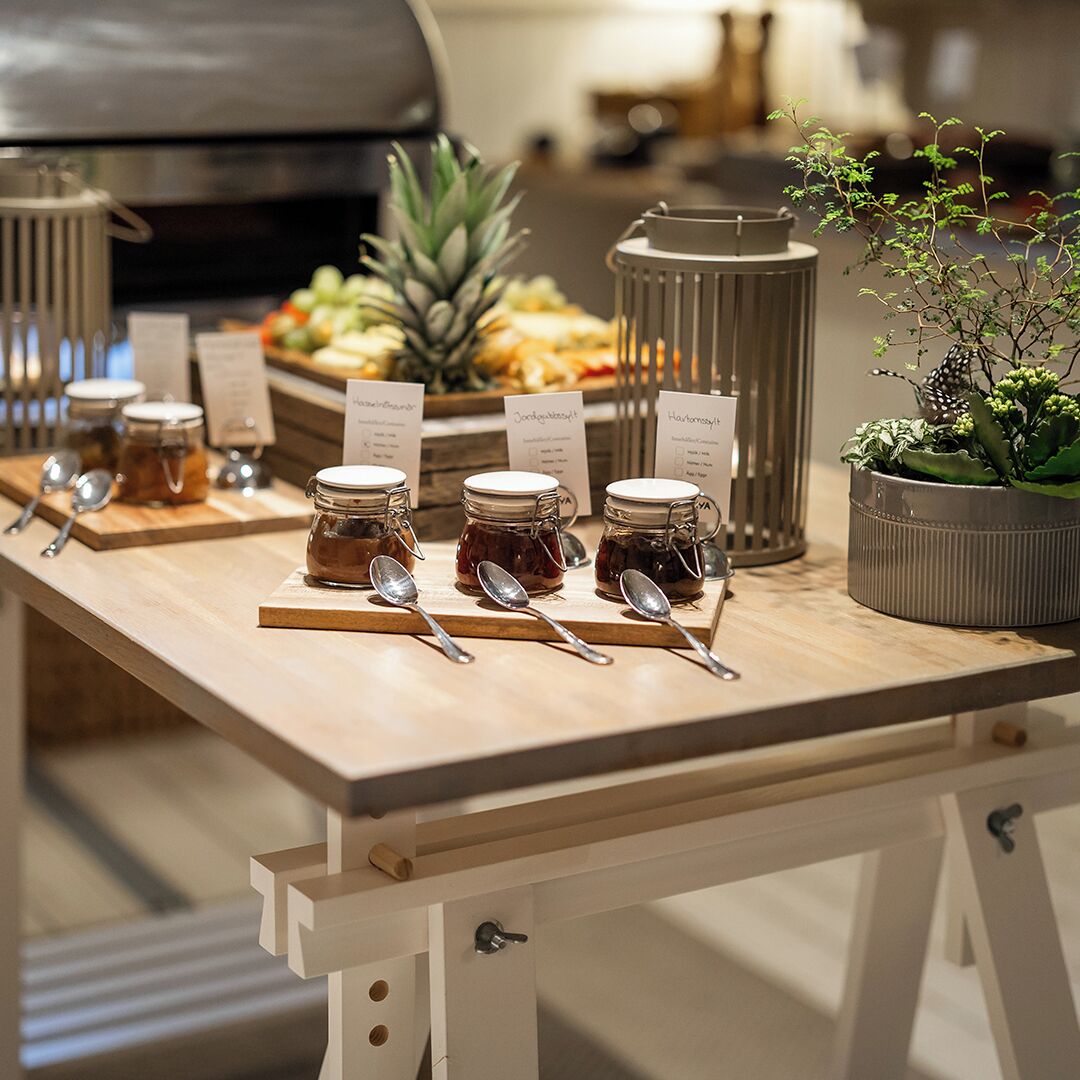 A wooden table with jars of condiments, spoons, and a potted plant.