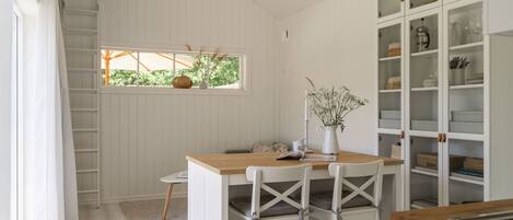 A kitchen with white cabinetry, a wooden dining table, and chairs. There is a window with a view of greenery outside.