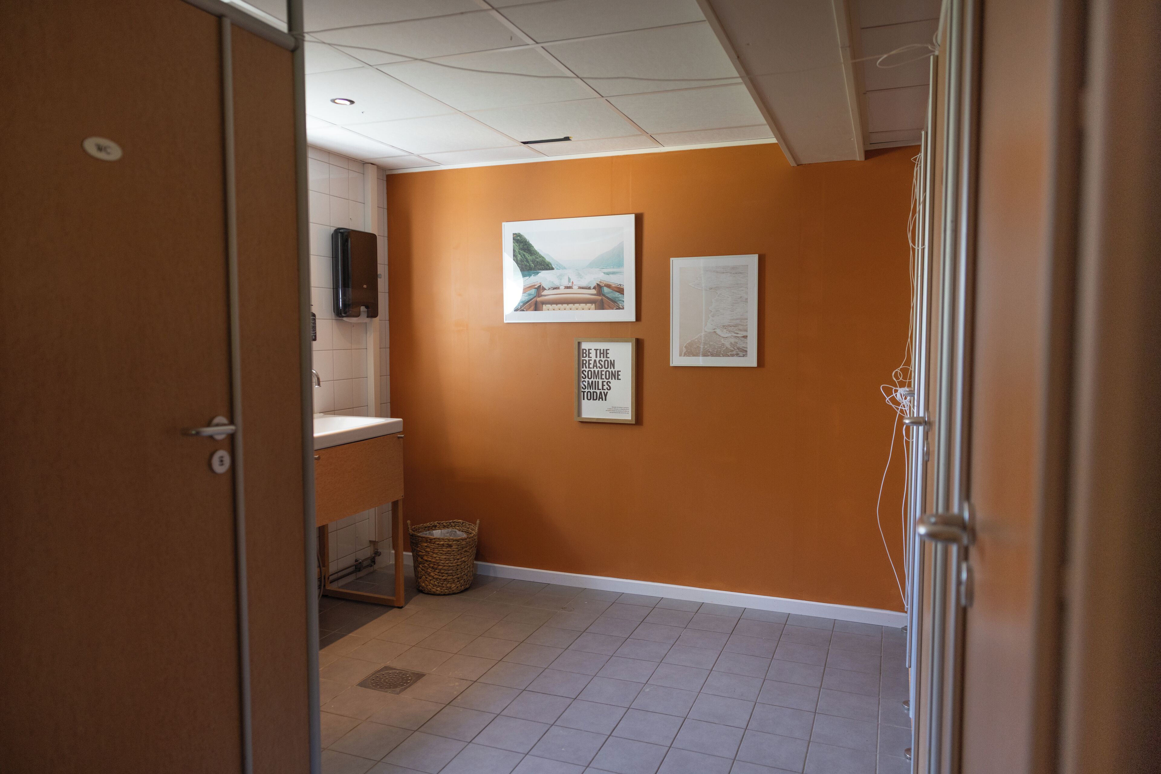Un pasillo con pared naranja, piso de baldosas y puerta que da a un baño. Hay cuadros enmarcados en la pared y una cesta en el suelo.