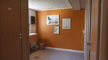 Un pasillo con pared naranja, piso de baldosas y puerta que da a un baño. Hay cuadros enmarcados en la pared y una cesta en el suelo.