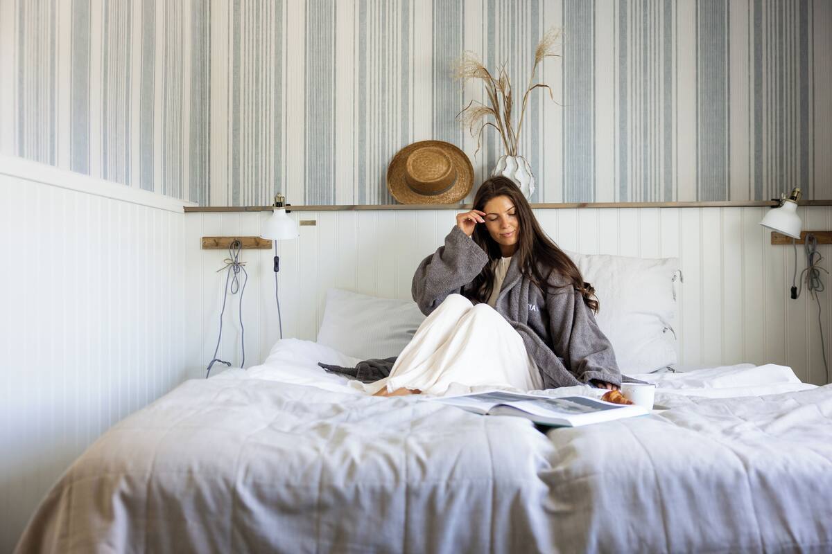 A person sitting on a bed with a book and a cup, in a room with striped wallpaper and a hat on the wall.