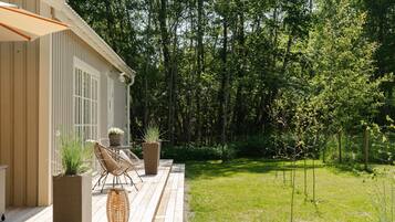 A wooden deck with wicker furniture and potted plants, overlooking a lush green garden and dense trees.