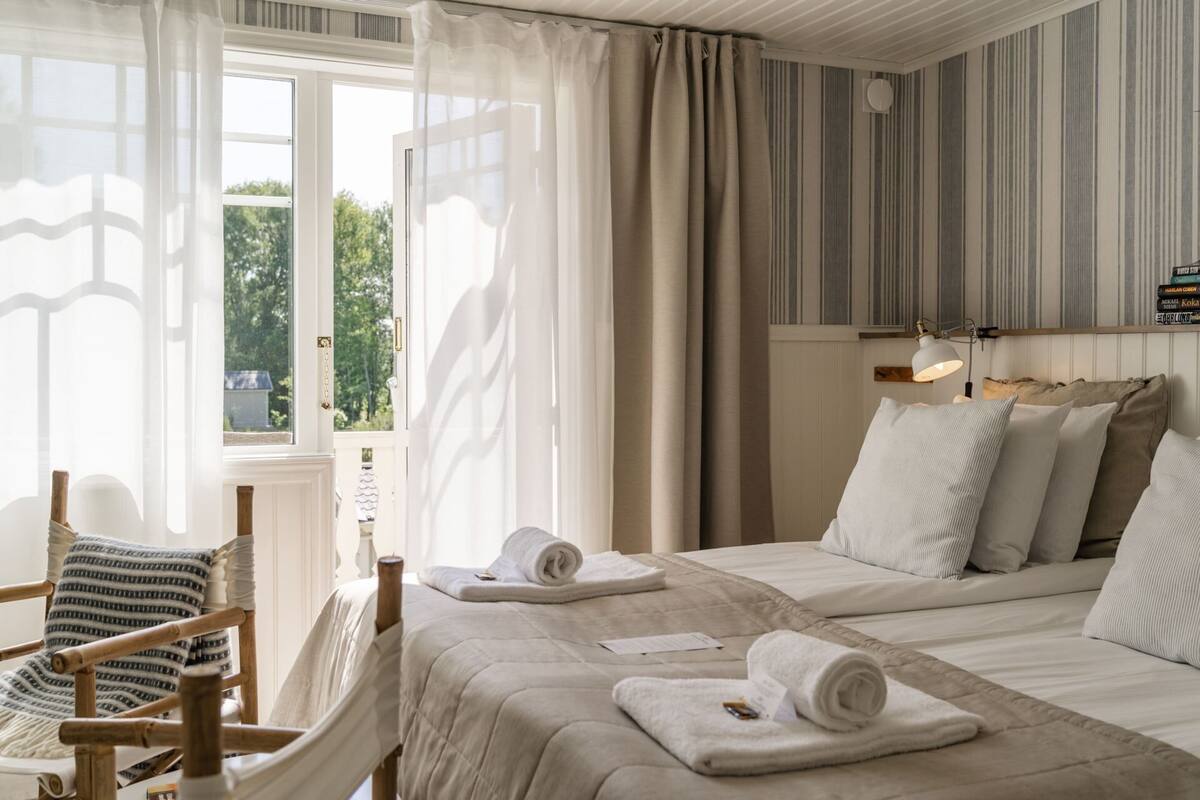 A hotel room with two beds, a wooden chair, and a window with sheer curtains.