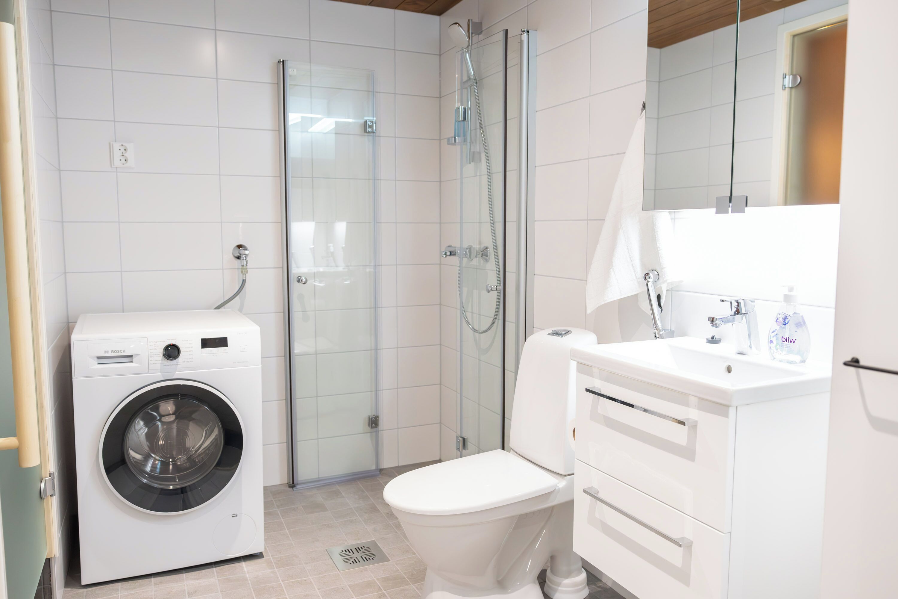 Een moderne badkamer met een Bosch wasmachine, een douche met glazen wanden en een witte wastafel met spiegel.