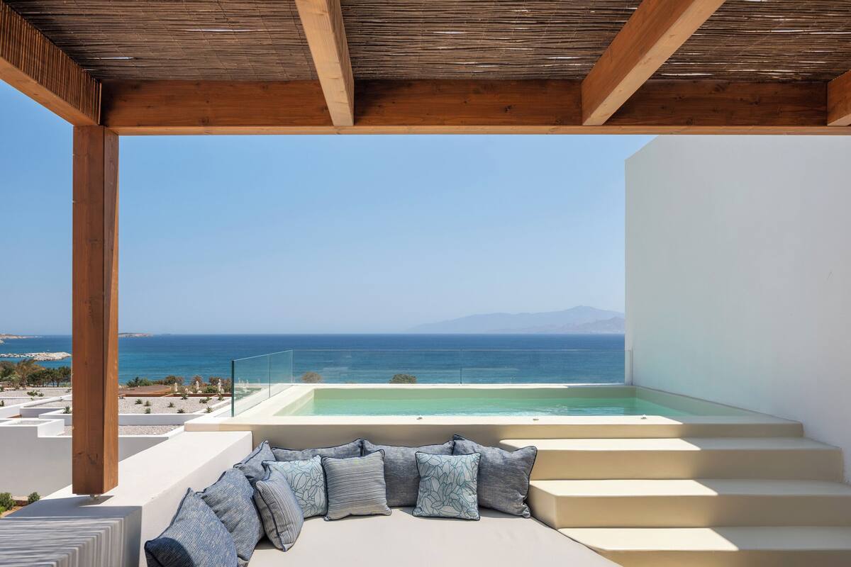 A balcony with a view of the sea and mountains, a pool, and a seating area with cushions.