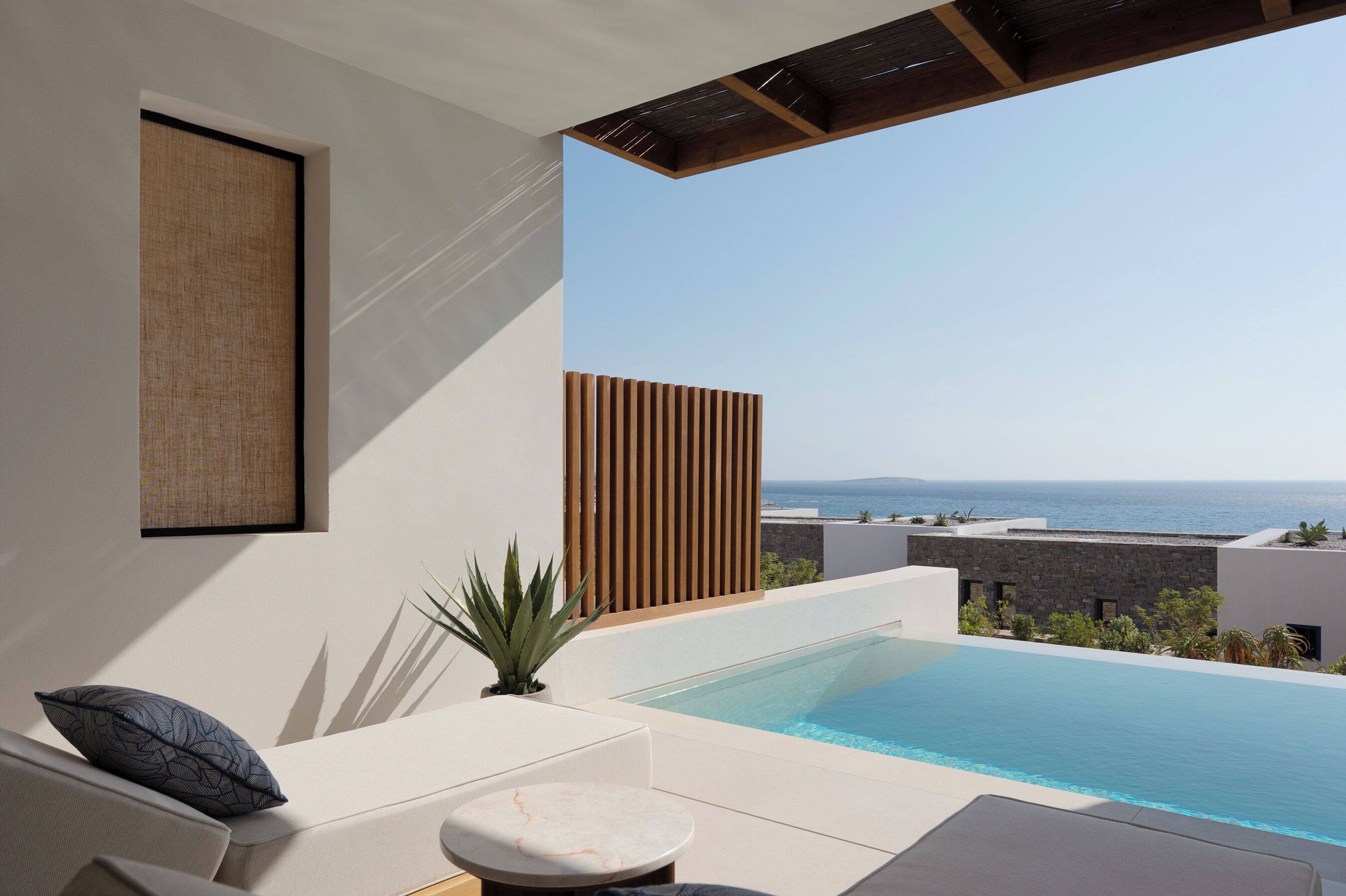 Area piscina moderna all'aperto con vista sul mare, zona relax con cuscini e un pannello divisorio in legno.