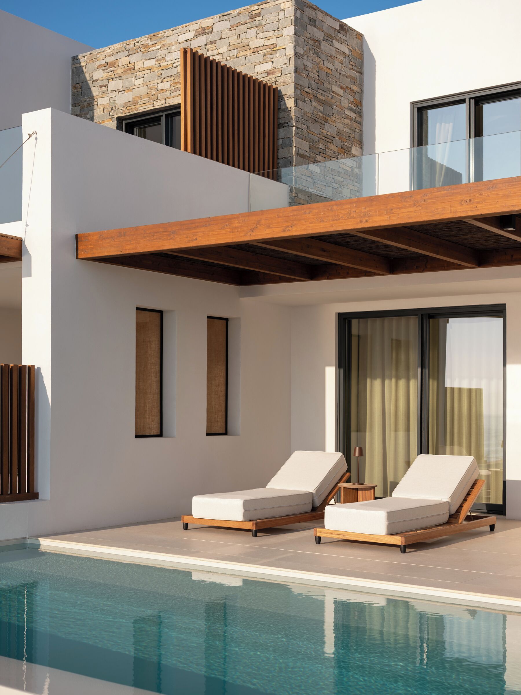 Un área de piscina moderna con dos sillones reclinables, fachada de piedra y barandilla de cristal.
