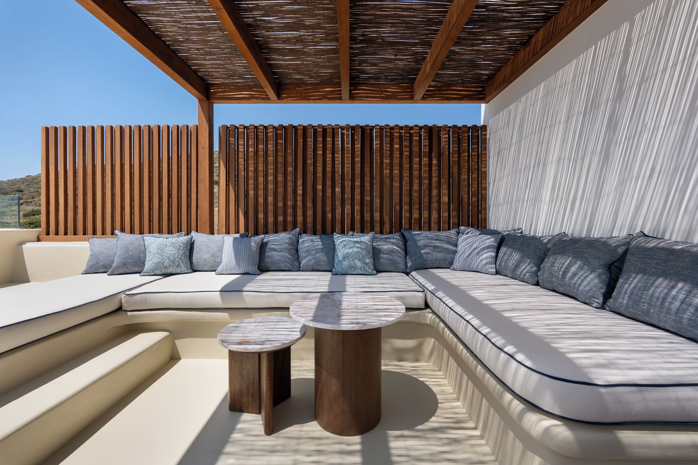 A modern outdoor seating area with a curved sofa, two round wooden tables, and a wooden pergola overhead.