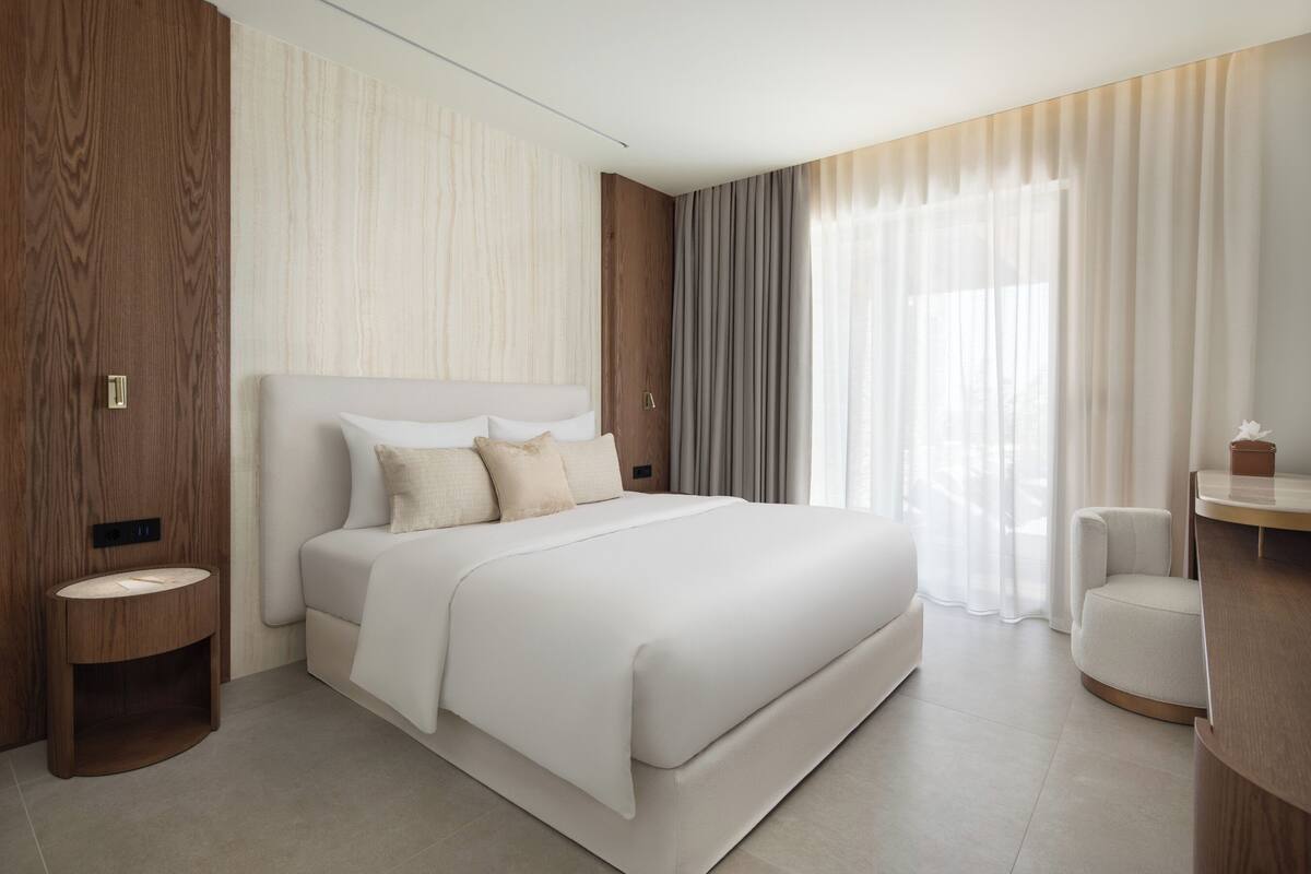 A modern hotel room with a large bed, a bedside table, a small round table, and a large window with curtains.