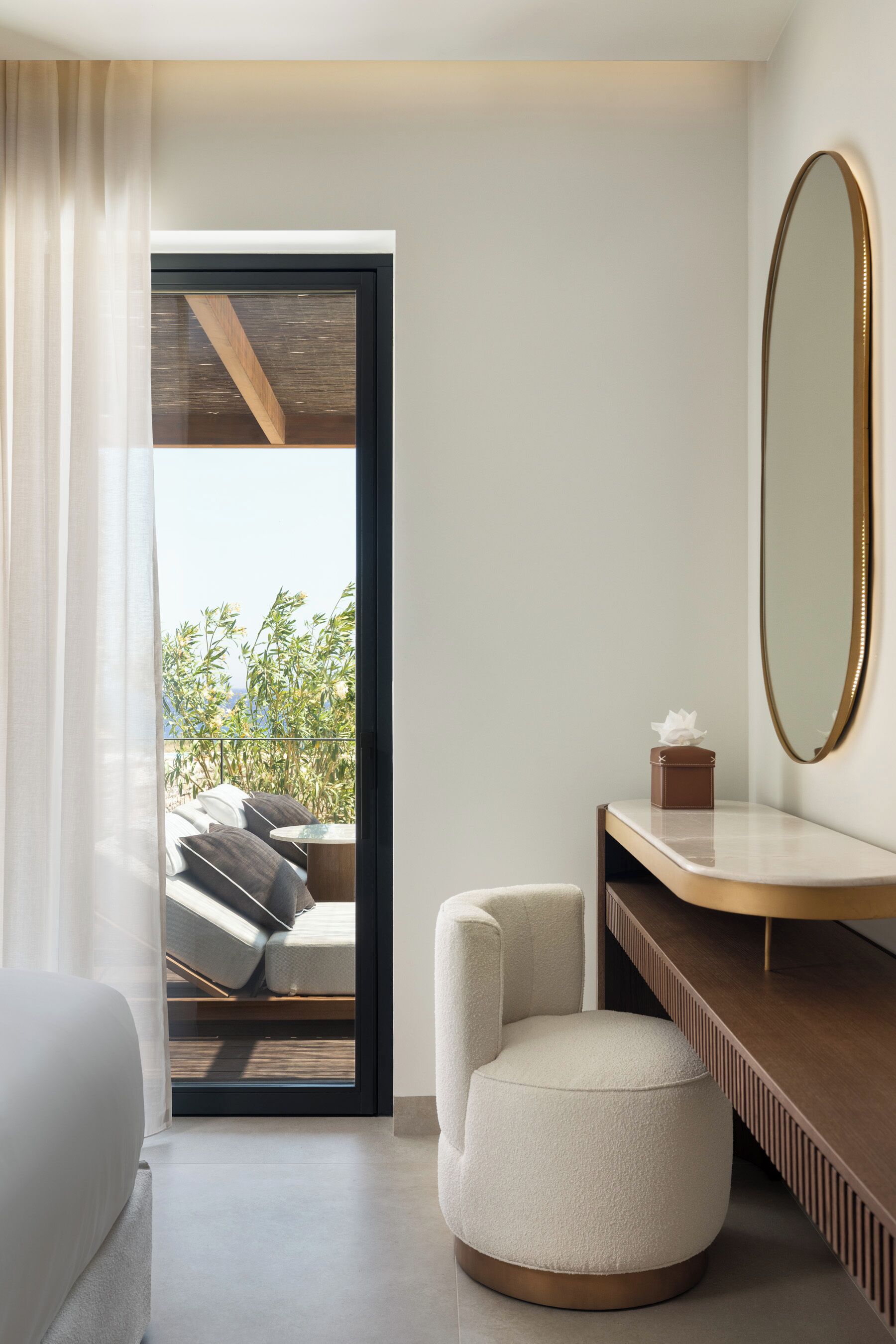 A modern hotel room with a balcony, a round ottoman, a bedside table with a tissue box, and a large mirror.