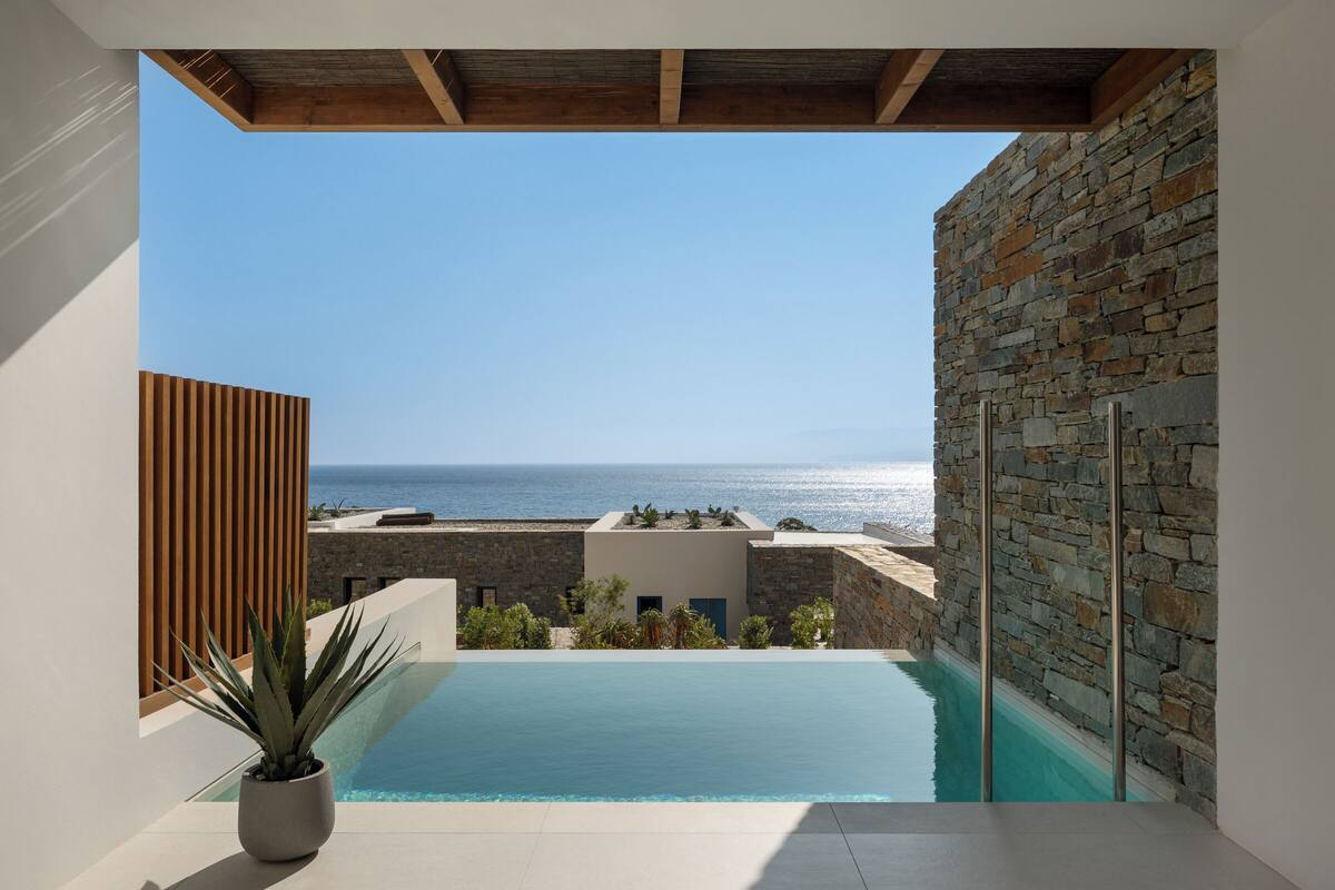 A modern pool area with a view of the sea, stone walls, and a wooden ceiling.