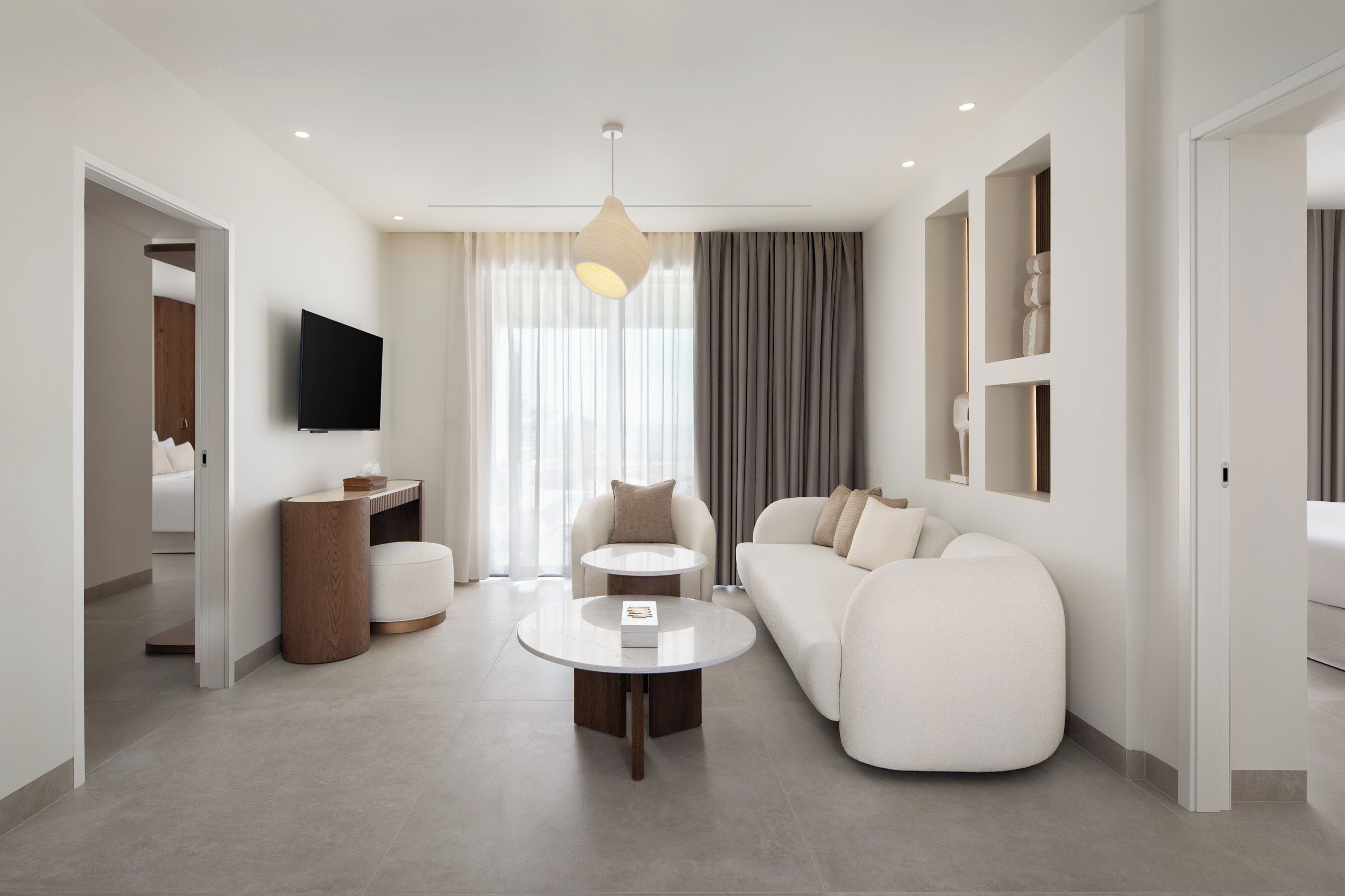 A modern hotel room with a sofa, a coffee table, a TV, and a large window with curtains.