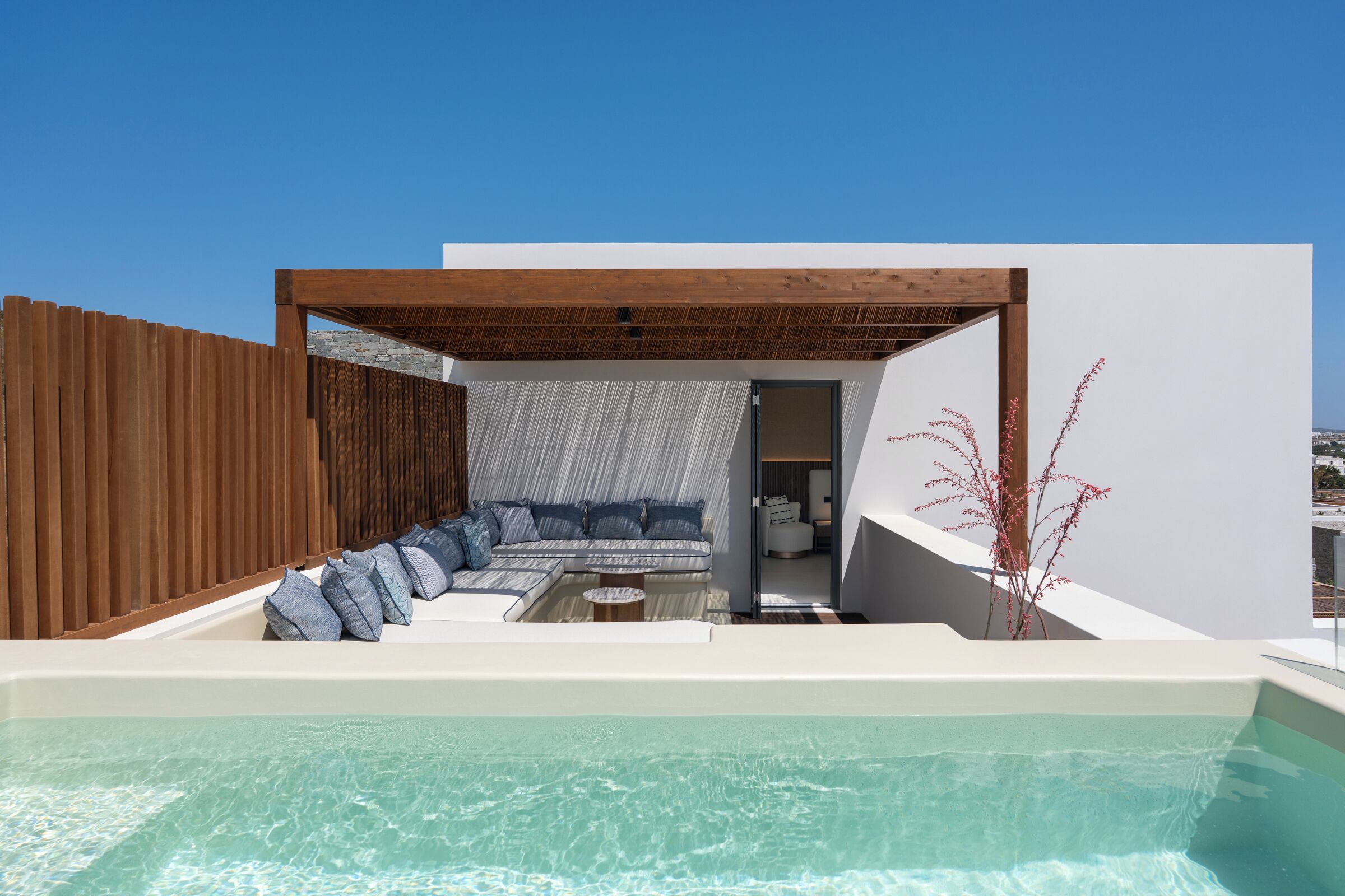 A modern outdoor pool area with a wooden pergola, a seating area with a sofa and cushions, and a view of the surrounding landscape.