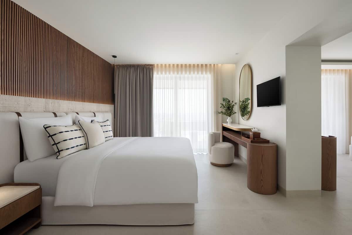 A modern hotel room with a large bed, a wooden headboard, a flat-screen TV, a bedside table, and a large window with curtains.