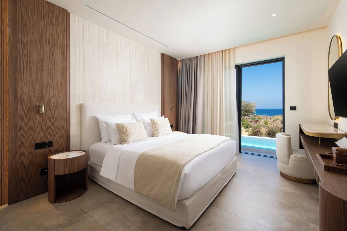 A modern bedroom with a large bed, a small bedside table, a desk with a chair, and a view of the sea through a glass door.