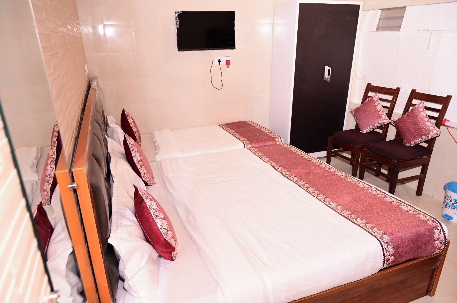 A hotel room with a bed, a TV mounted on the wall, a door, and two wooden chairs with cushions.