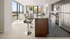A modern kitchen with stainless steel appliances, a central island with bar stools, and large windows offering a city view. - 10 min drive to Capitol 2 BR | Crystal City (Arlington)
