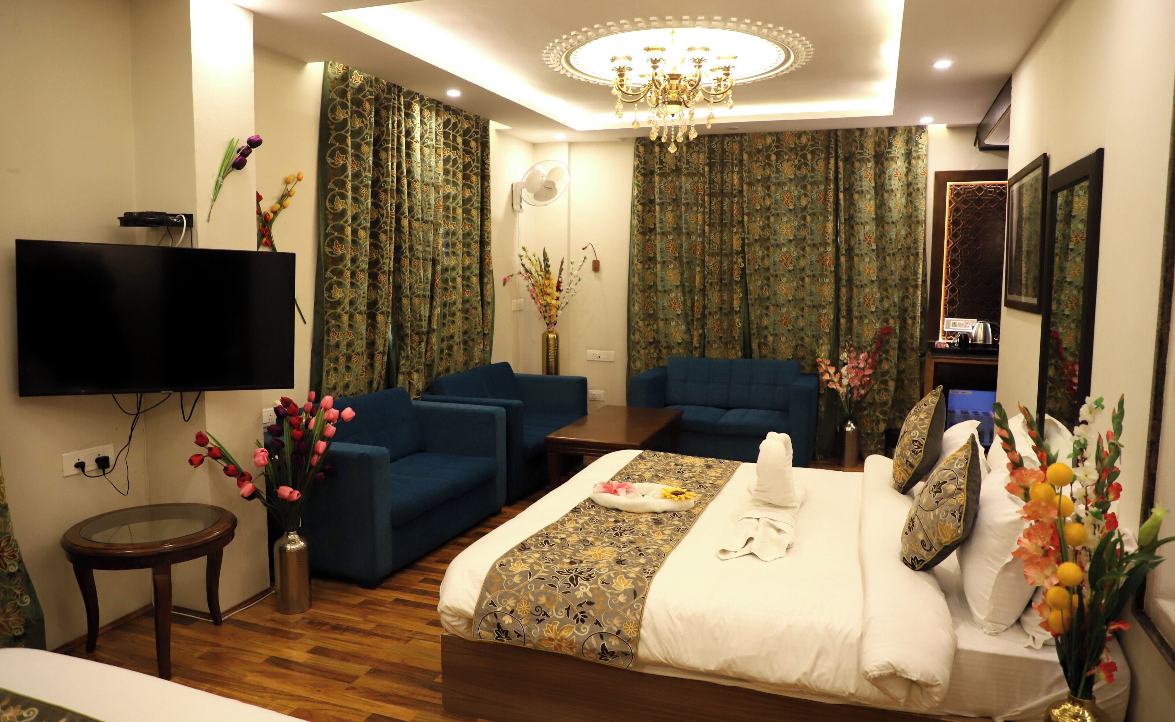 A hotel room with a bed, two blue armchairs, a TV, a chandelier, and floral decorations.