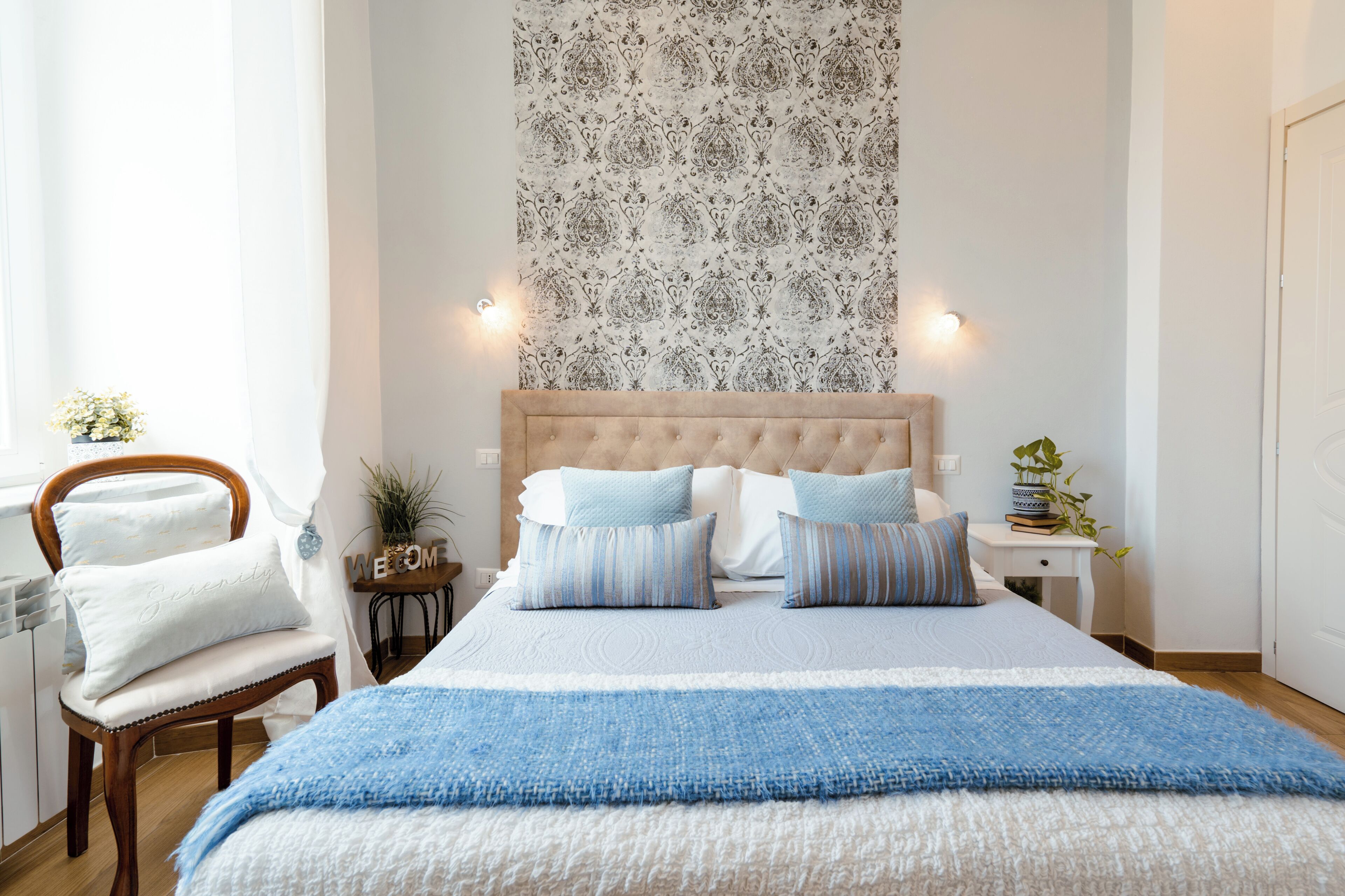 Ein Schlafzimmer mit einem großen Bett, einem Holzstuhl, einem Nachttisch und einer Wand mit ornamentalem Wandpapier.
