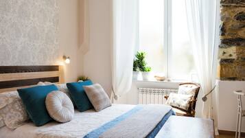 Un dormitorio con una cama, almohadas, una mesita de noche, una silla, una ventana con cortinas y una planta.