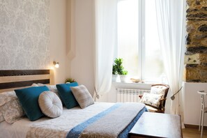 Ett sovrum med en säng, kuddar, ett sängbord, en stol, ett fönster med gardiner och en växt.