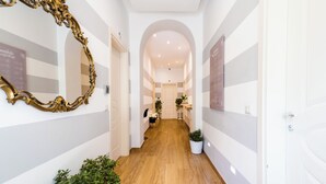 A hallway with a wooden floor, white walls, and a large ornate mirror on the left. There are potted plants along the sides and a door at the end. - The Railway 74 (La Spezia)