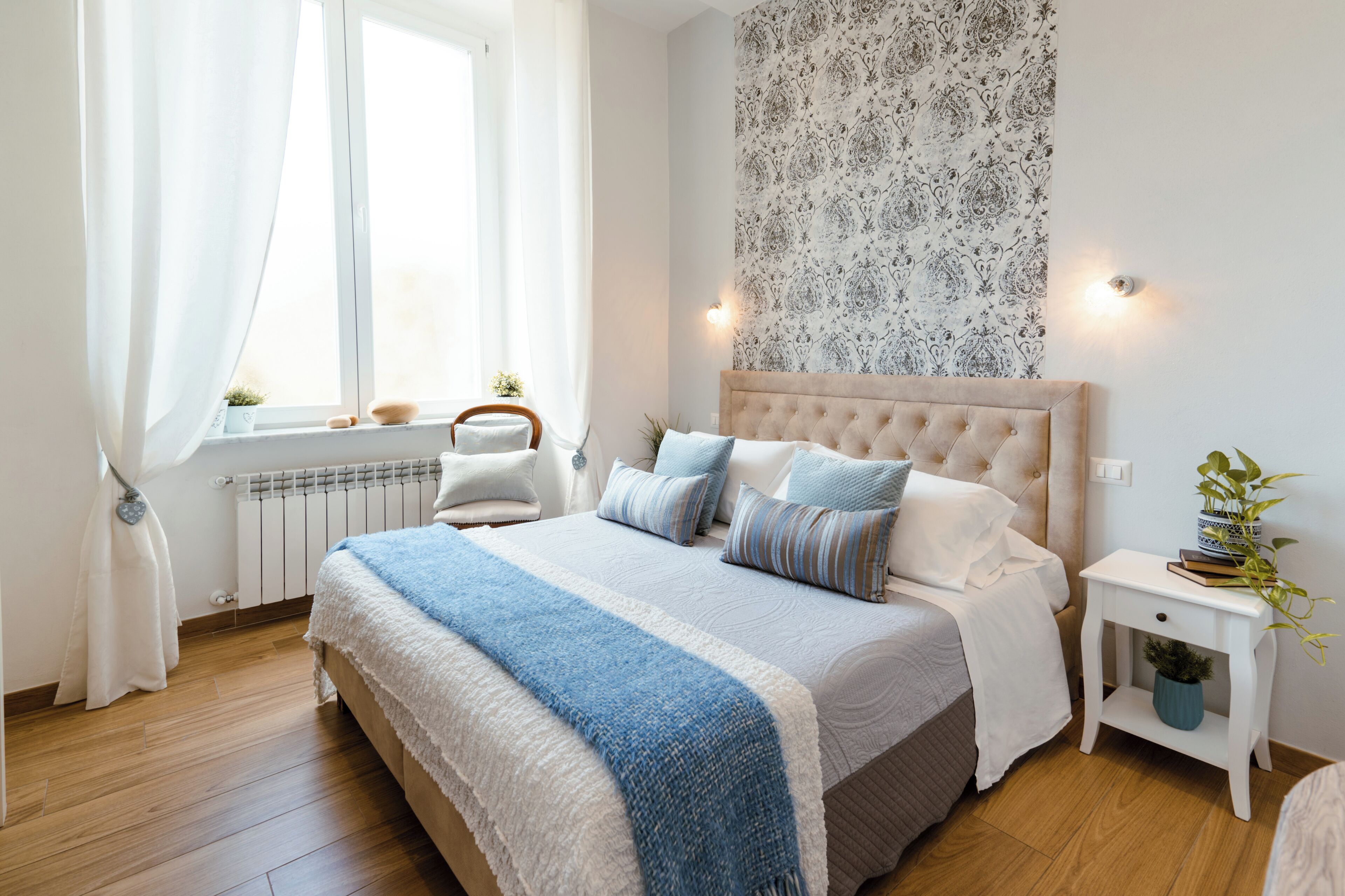 Una camera da letto con un letto grande, un termosifone, una finestra con tende e un comodino con una pianta, e una parete decorata.