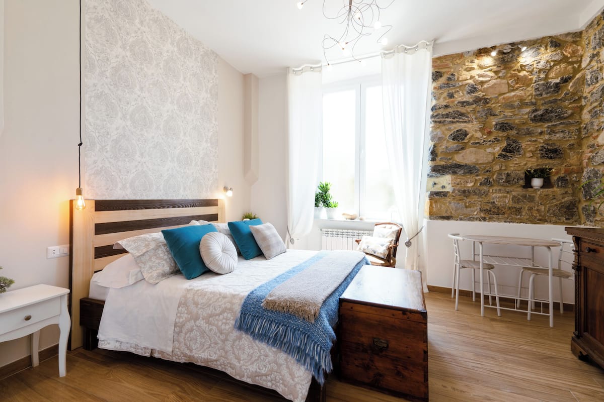 Une chambre avec un lit en bois, une table de chevet blanche, un bureau avec des chaises et un mur en pierre.