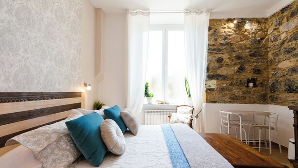 A bedroom with a wooden headboard, a large bed with patterned bedding, a stone wall, a window with curtains, and a small table with chairs. - The Railway 74 (La Spezia)