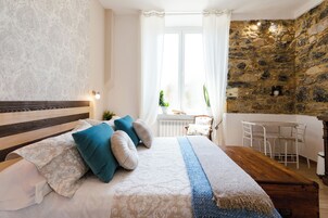 Un dormitorio con cabecera de madera, una cama grande con ropa de cama estampada, una pared de piedra, una ventana con cortinas y una pequeña mesa con sillas.