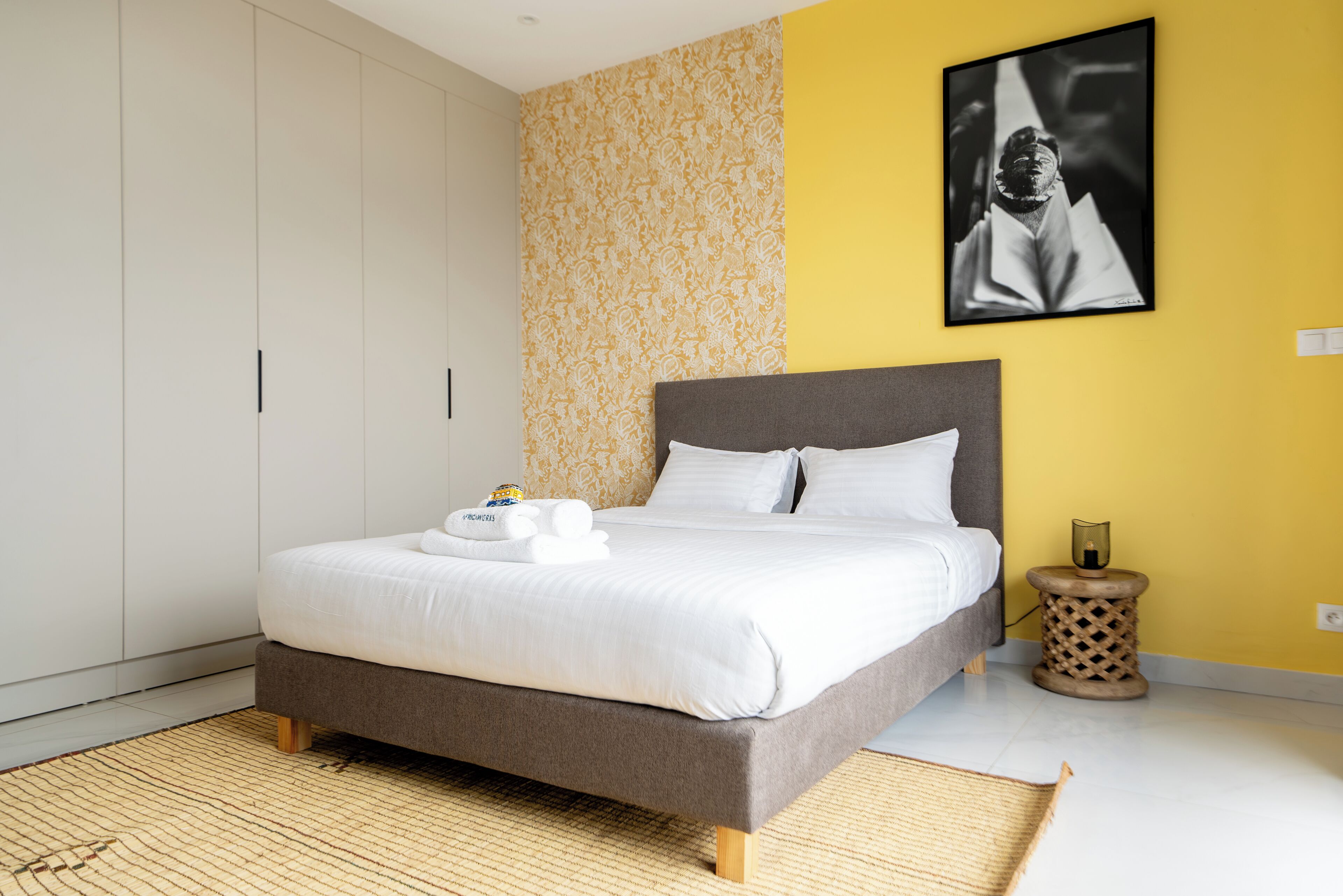 A neatly made bed with white linens and a gray headboard in a modern bedroom with a yellow accent wall and a framed picture.