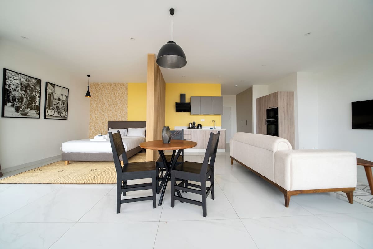 A modern living room with a sofa, dining table, and chairs. There is a kitchen area with cabinets and a countertop.