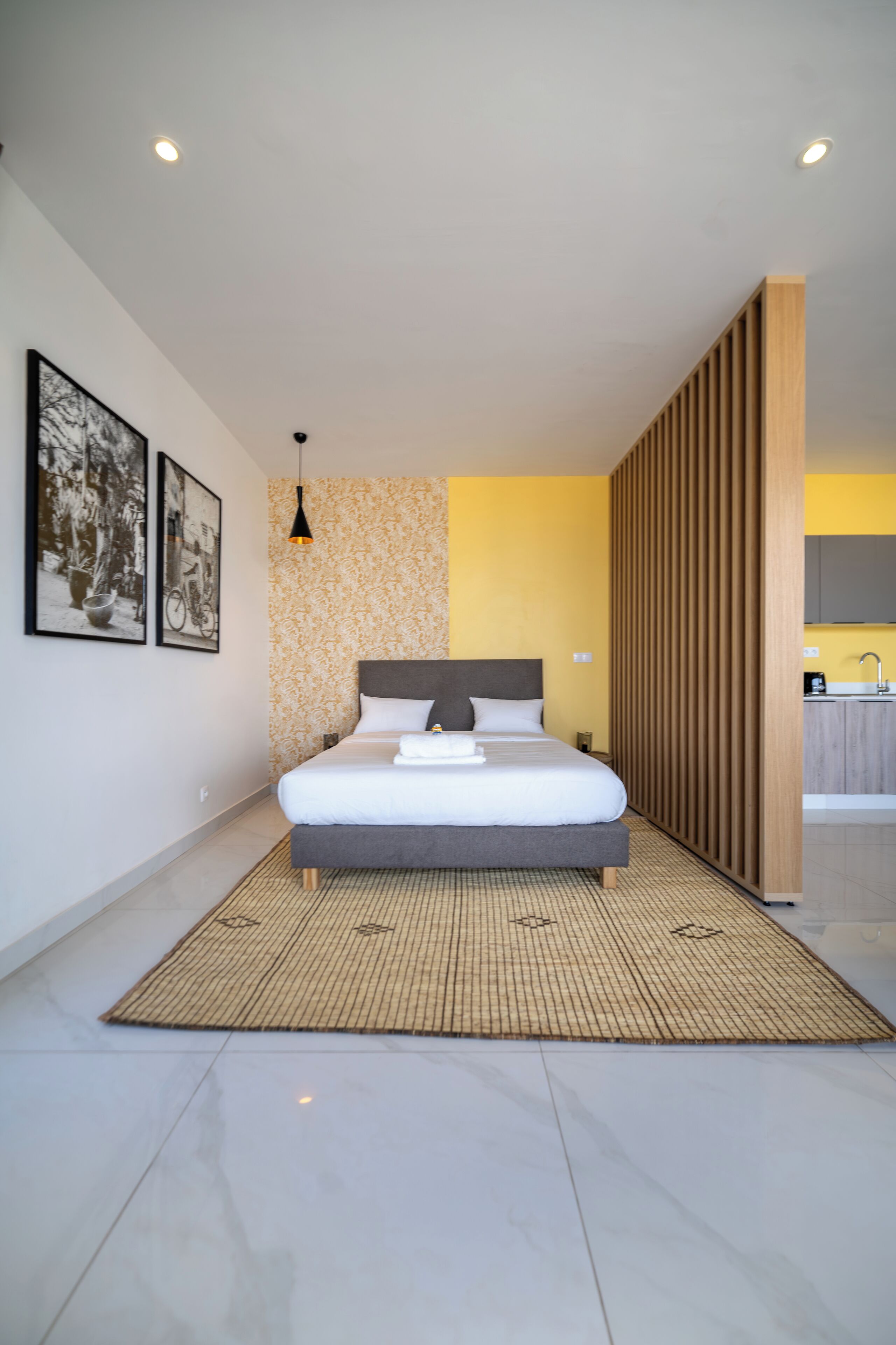 A modern bedroom with a large bed, a patterned wall, and a kitchen area visible through an open door.