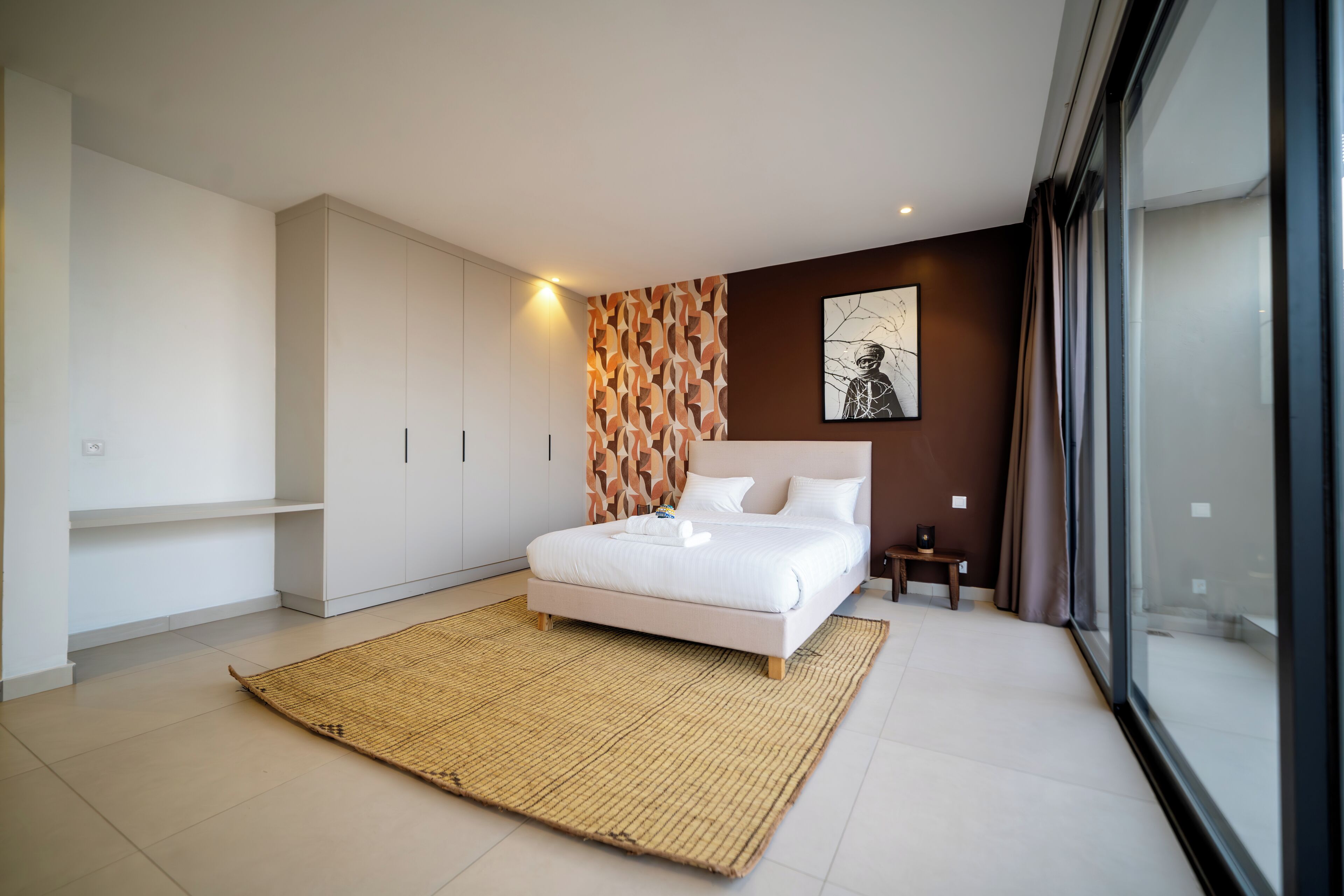Un dormitorio moderno con una cama grande, un panel decorativo en la pared, una mesita de noche pequeña y un ventanal amplio.