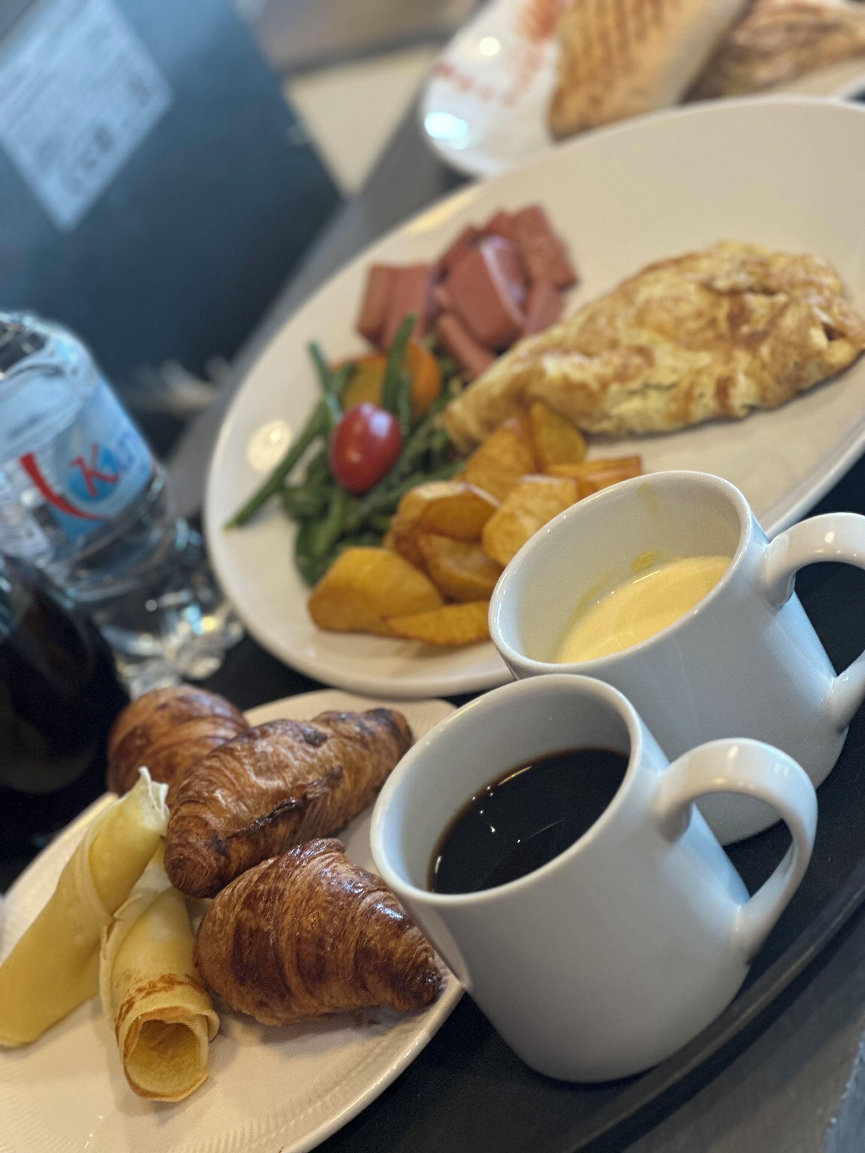 Una bandeja con croissants, un plato de comida que incluye un croissant, papas fritas, una ensalada y un sándwich, junto con dos tazas de café y una botella de agua.